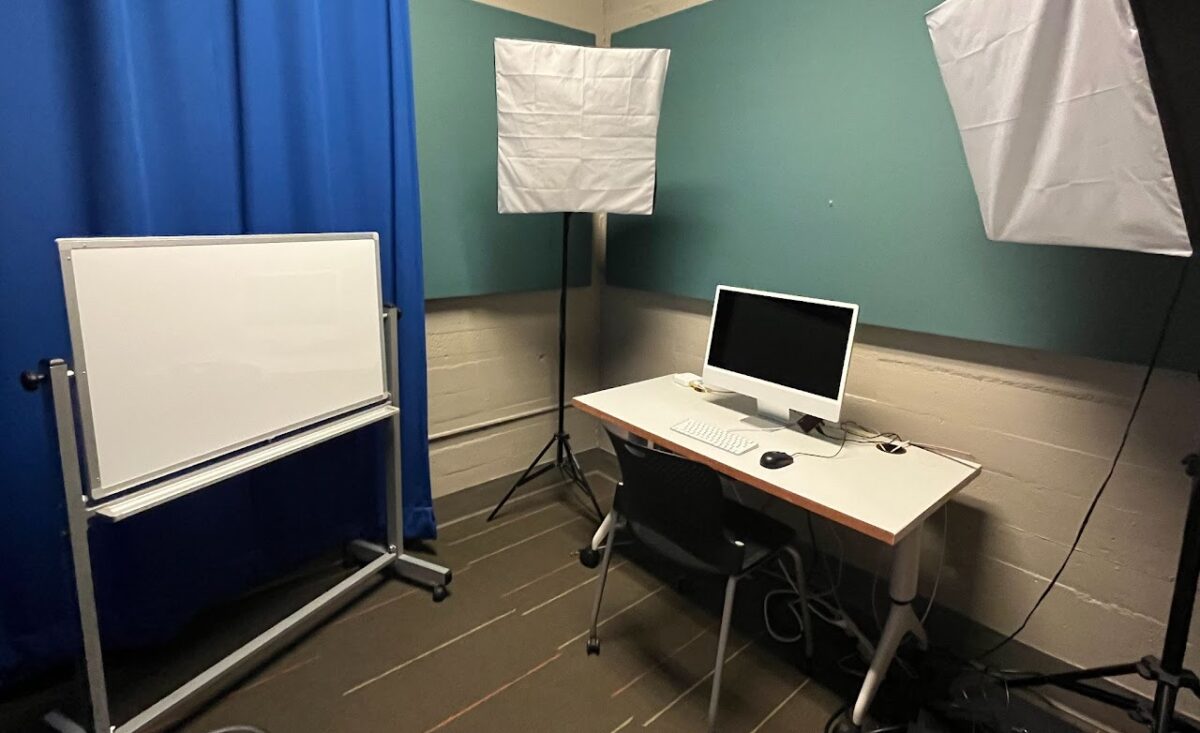 Vault Recording Studio with two studio lights, a desk with a Mac Computer,  soundproofing, a blue backdrop, and a mini-whiteboard